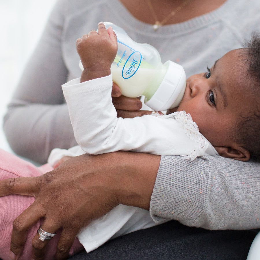 A baby drinking from a milk bottle while being held by an adult. The baby is wearing a white long-sleeve shirt and pink pants. The adult is wearing a gray sweater, and a wedding ring is visible on their hand.