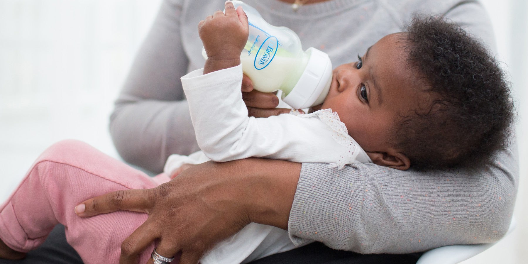 An infant lying in someone's arms, wearing a white top and pink pants, is drinking from a baby bottle. The person holding the baby is wearing a gray sweater.