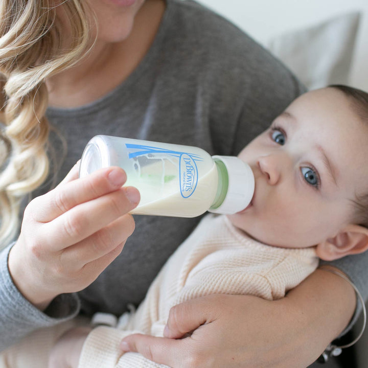 A Dr. Brown's narrow glass baby bottle with a white cap and a light green translucent collar, filled with milk, being held by a woman and offered to a baby wearing a cream knit outfit.