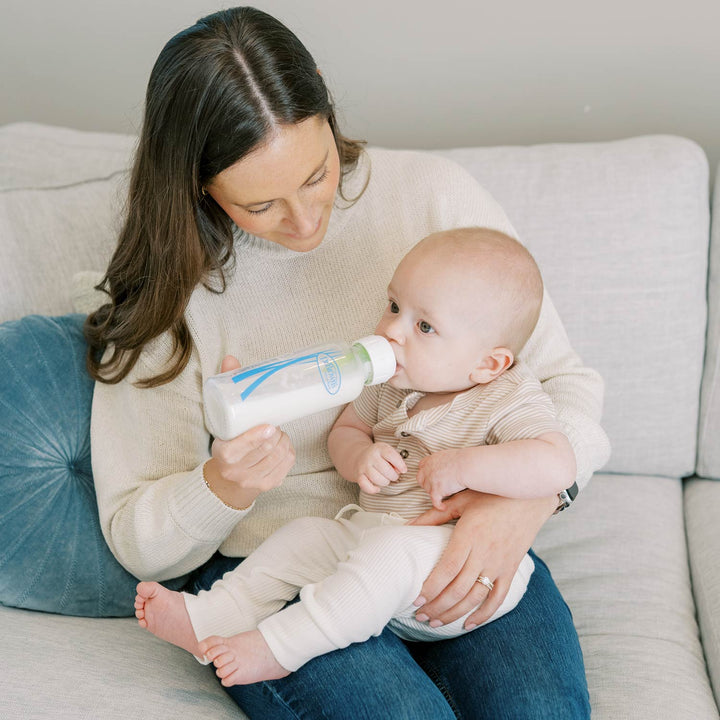 A woman sitting on a light gray couch holds a baby wearing a beige striped long-sleeve shirt and cream pants while the woman feeds the baby from a narrow glass Dr. Brown's baby bottle.