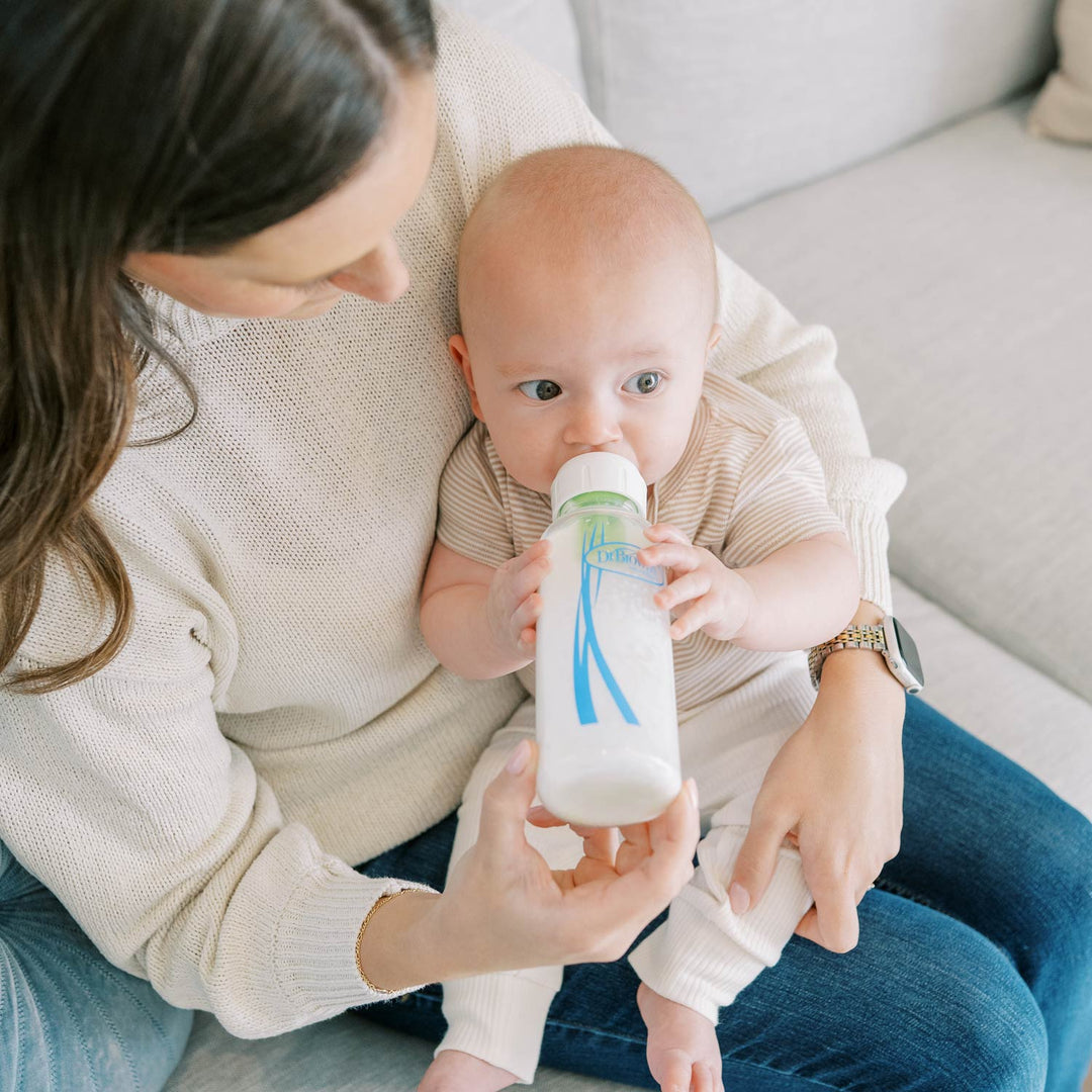 A Dr. Brown's narrow glass baby bottle with a white and green cap and blue graphic, being held to a baby's mouth while a woman in a cream knit sweater sits on a light gray sofa.