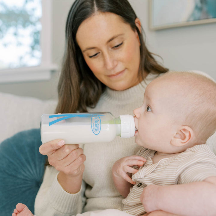A woman holding a baby while offering a glass baby bottle with a blue logo. The baby is wearing a beige striped outfit, and the scene is set in a softly lit indoor environment.