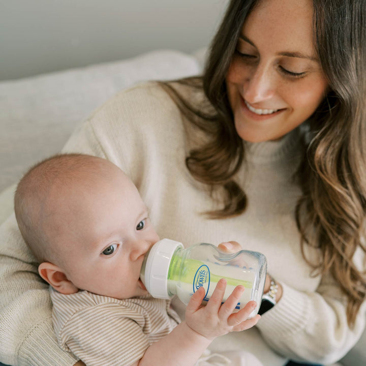 A baby drinking from a clear baby bottle with a white cap and green inner liner, held by a smiling woman in a cream sweater.