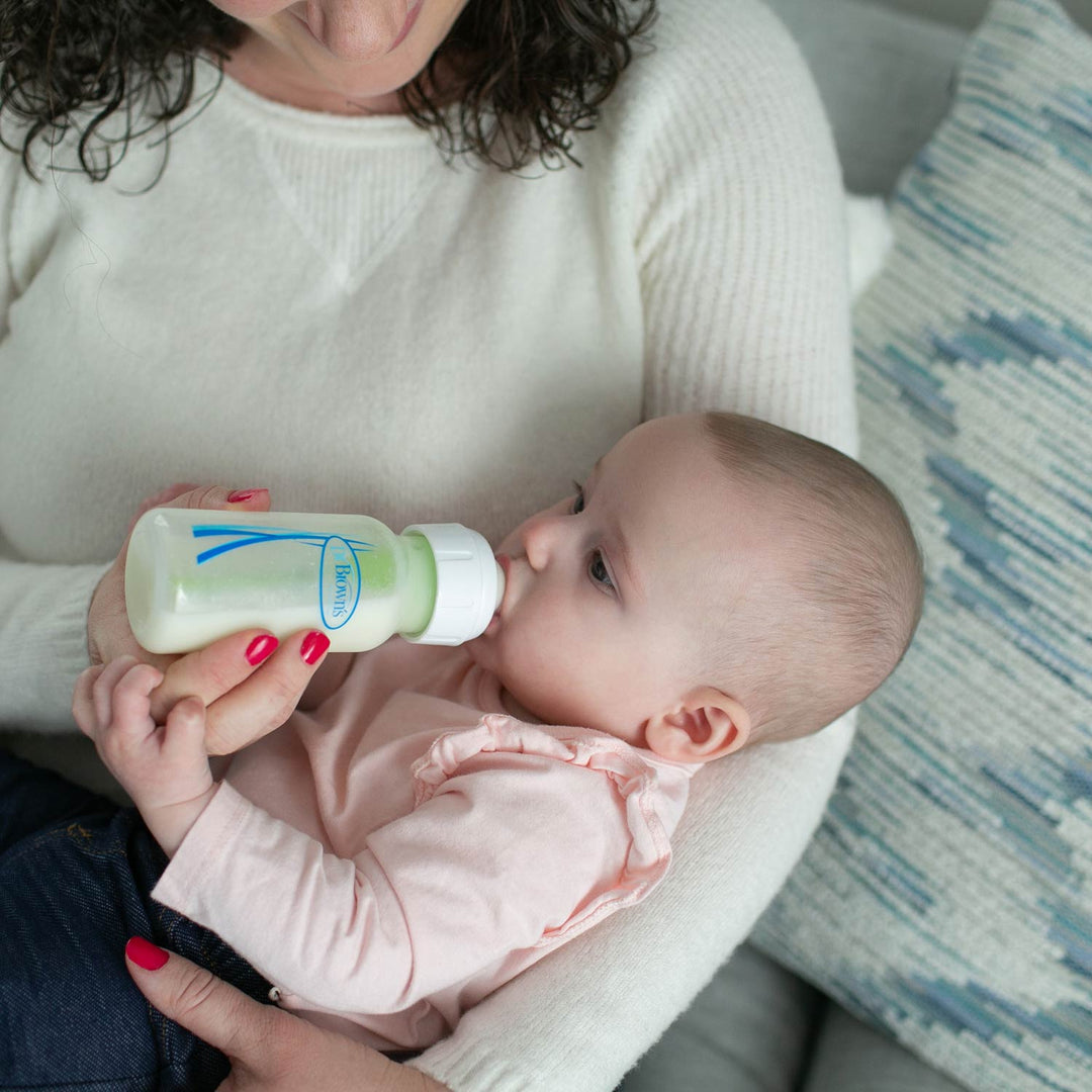 A baby wearing a light pink long-sleeve shirt is being bottle-fed by a woman in a cream-colored knit sweater. The baby is holding a white bottle with blue and green branding.