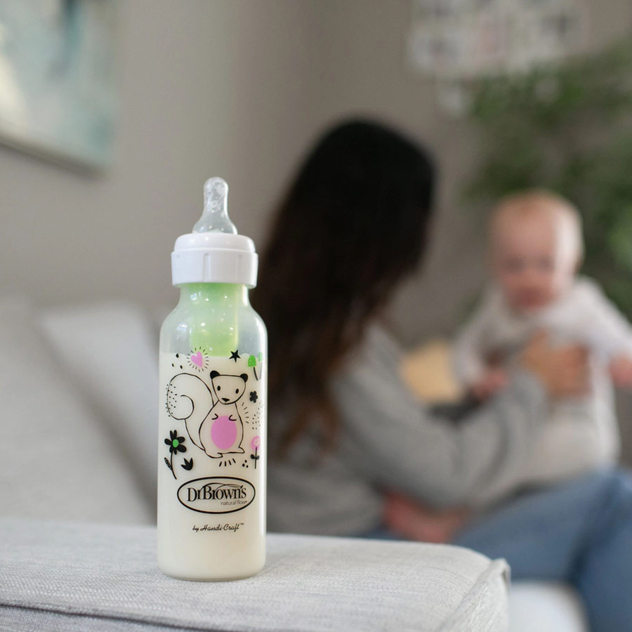 A baby bottle with a cute animal design placed on a soft surface. In the blurred background, a person is holding a baby in a cozy room.
