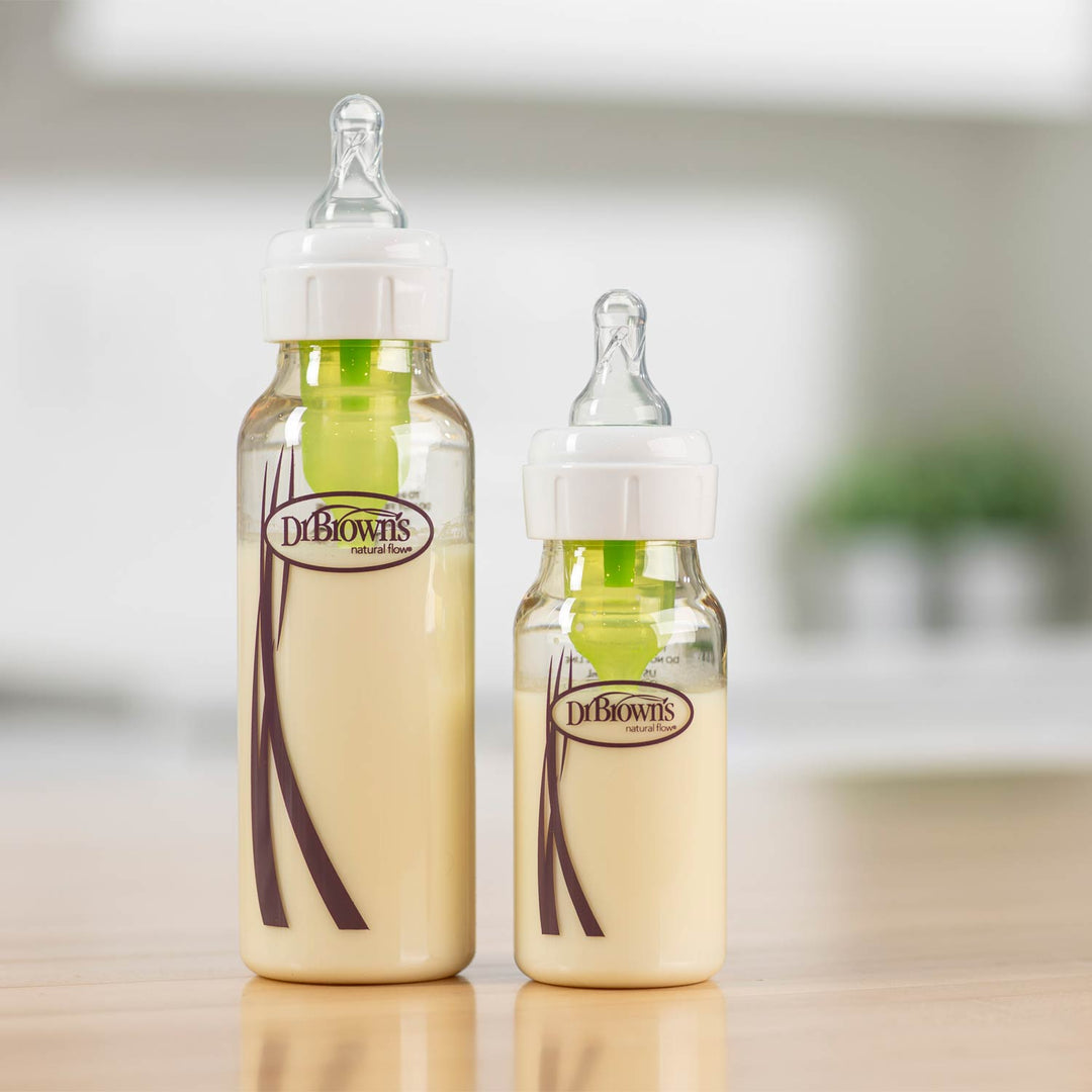 Two Dr. Brown's baby bottles, one large and one small, filled with milk and standing on a light wooden surface with a blurred kitchen background.