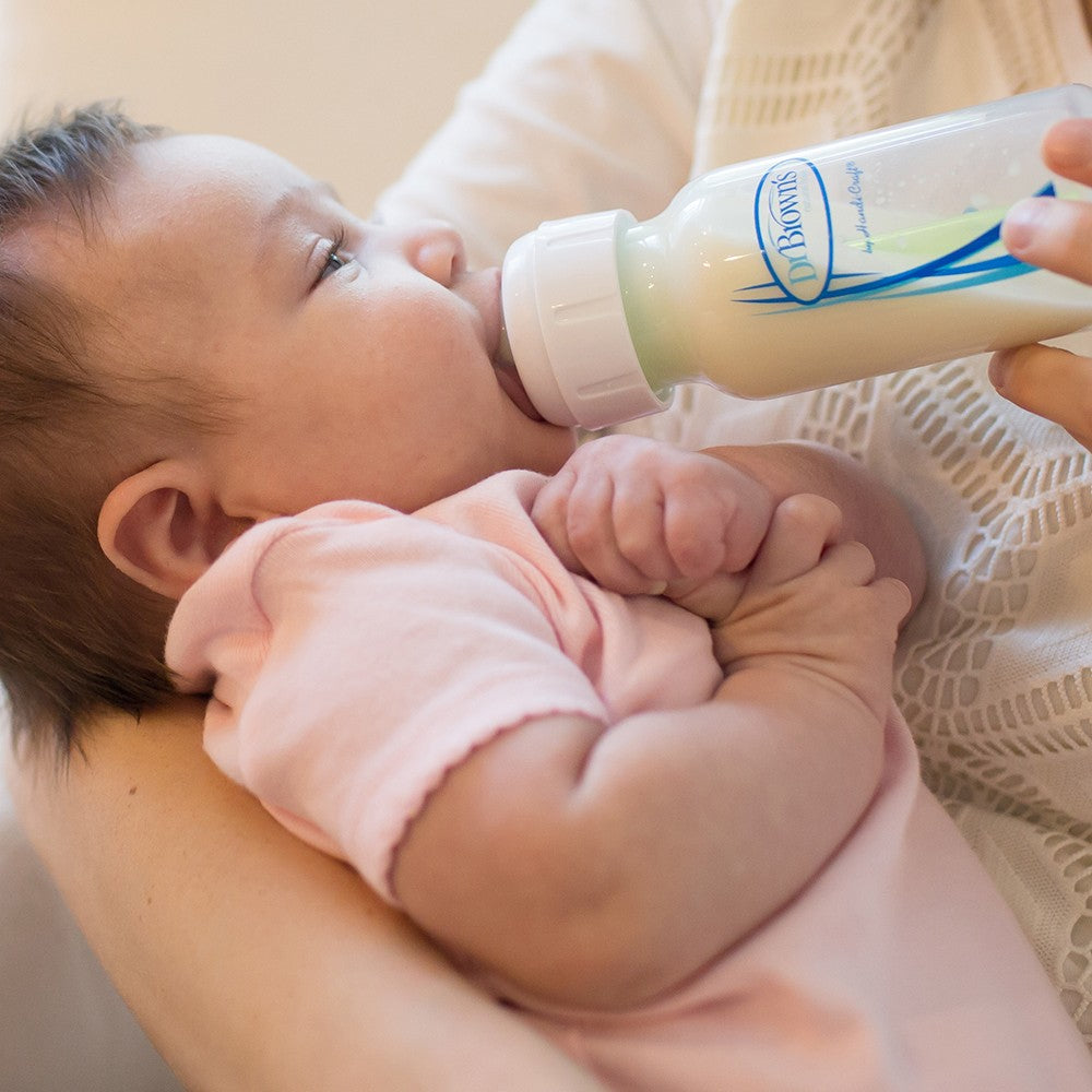 A baby in a pink outfit is being held and fed with a baby bottle. The person holding the baby is wearing a light-colored garment. The baby is looking up at the bottle.