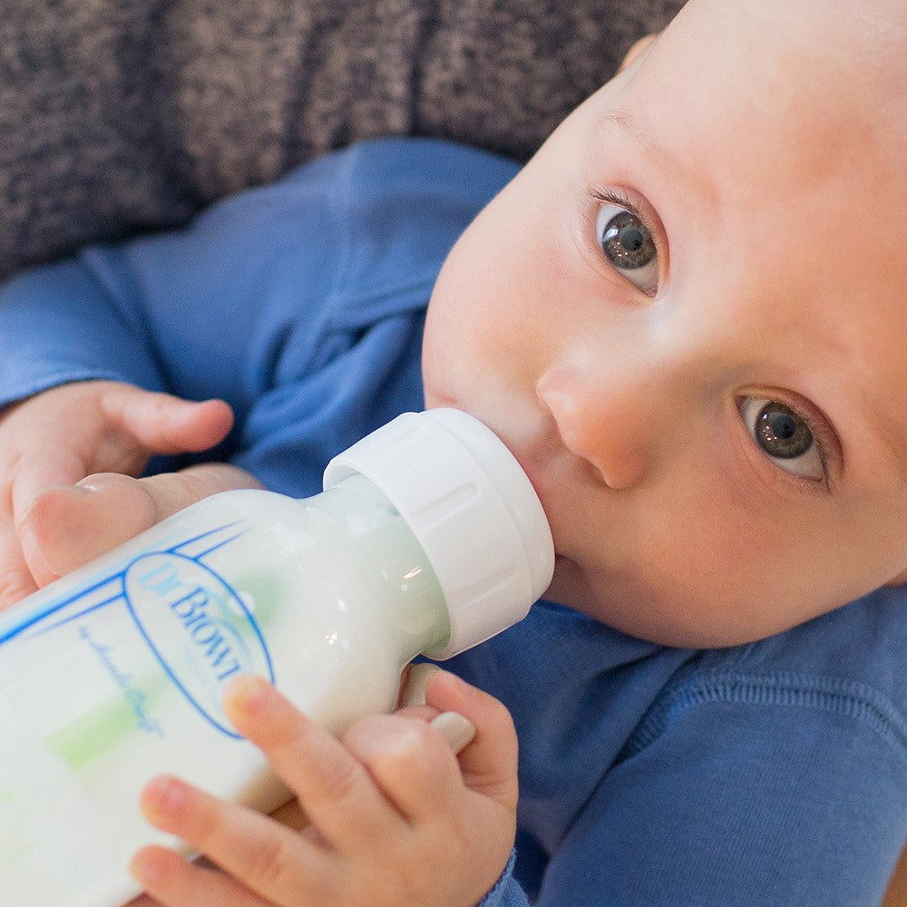 A baby in a blue outfit is drinking from a Dr. Brown's baby bottle. The baby has light skin and blue eyes and is lying down while holding the bottle with both hands. The bottle appears to be filled with milk or formula.