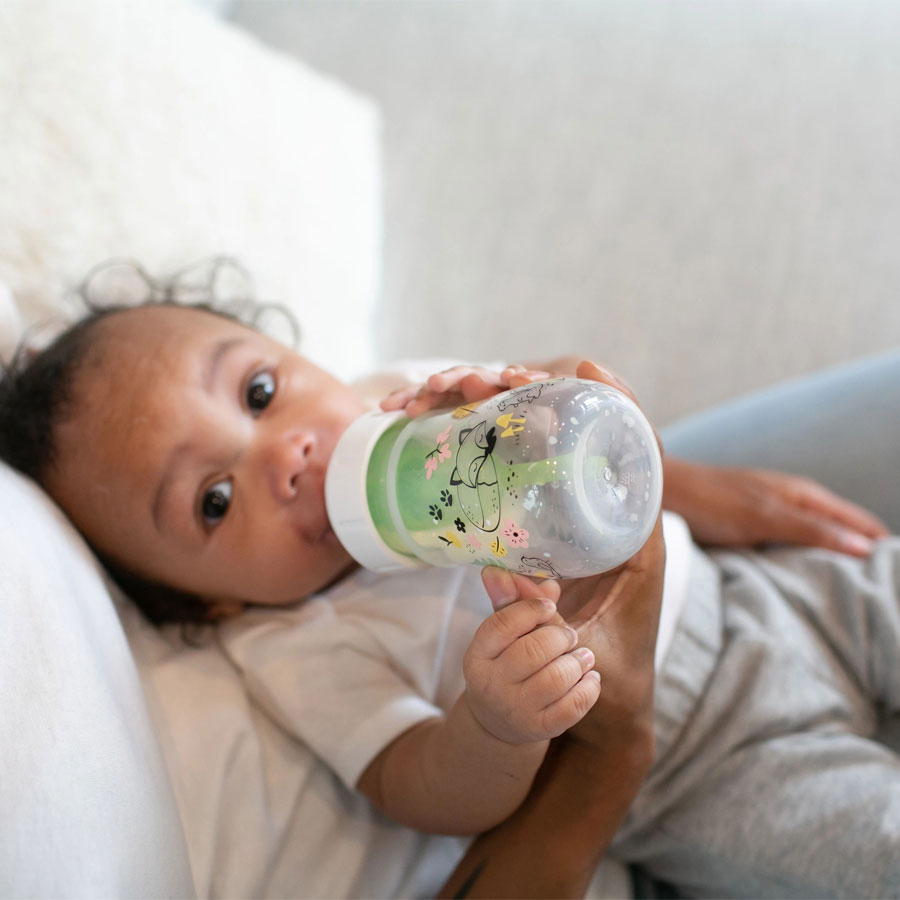A baby lying on a person's lap is drinking from a decorated baby bottle. The baby is wearing a white shirt and gray pants, and the background is soft and blurred.