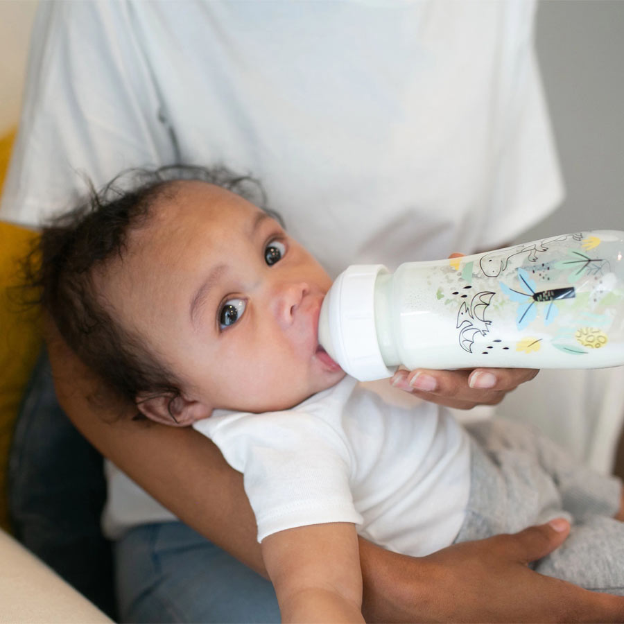 A baby drinking milk from a bottle held by an adult. The baby is wearing a white shirt and gray pants, resting on the adult's lap. The adult is wearing a white shirt and sitting on a yellow chair.