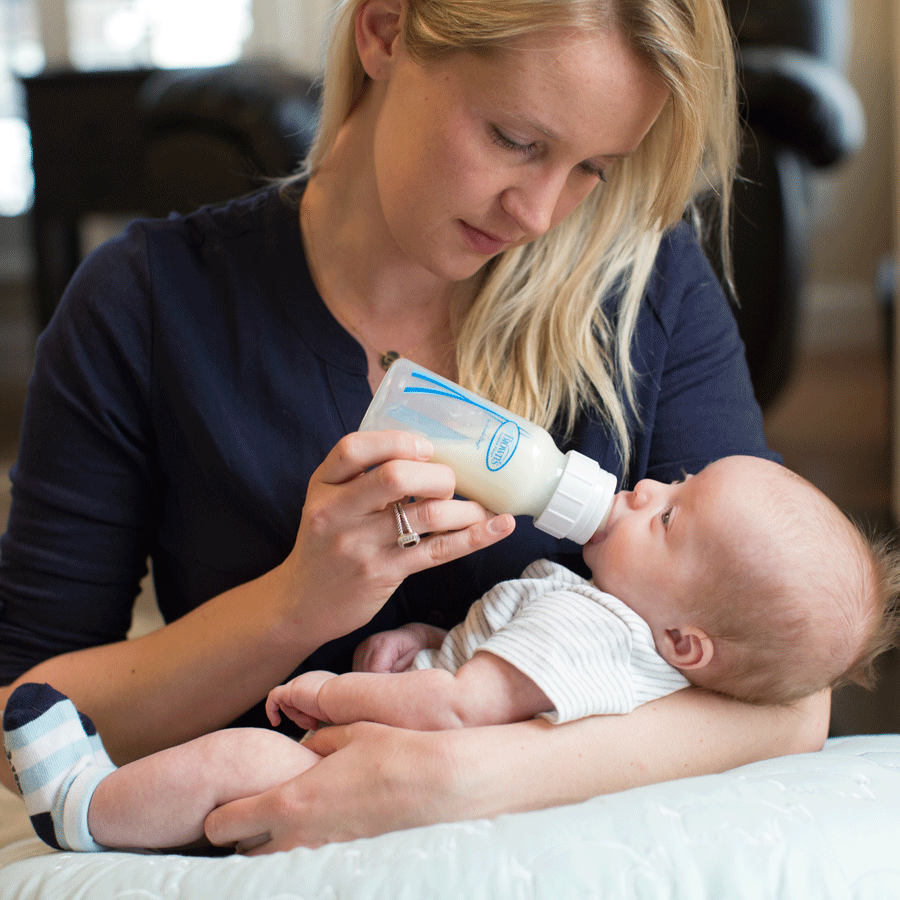 A woman with blonde hair is sitting and bottle-feeding a baby dressed in a white outfit. The baby is cradled in her arms, and she is looking at the baby attentively. The setting is a cozy indoor space with soft lighting.