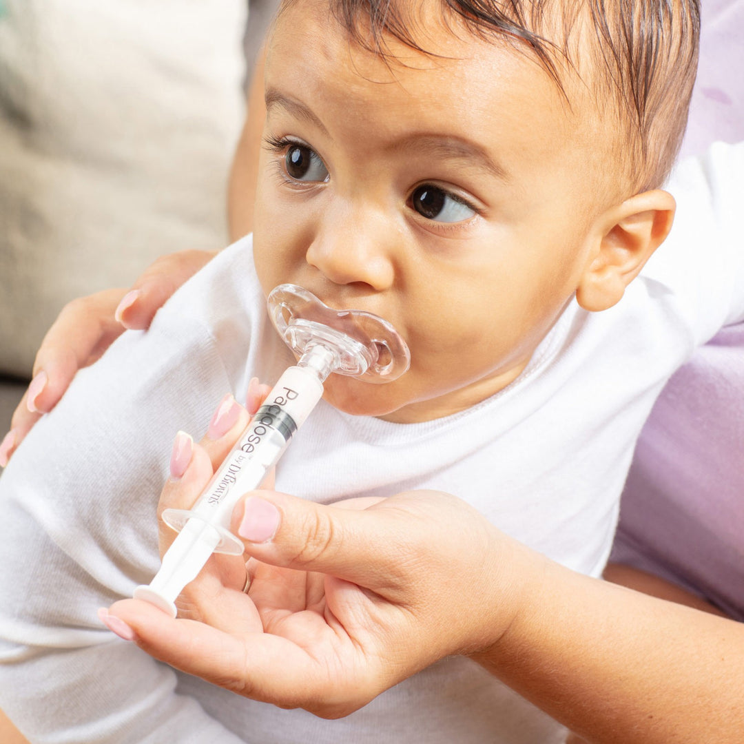 A baby is being fed medicine with a pacifier-shaped syringe. An adult's hands are holding the syringe and supporting the baby, who is wearing a white shirt.