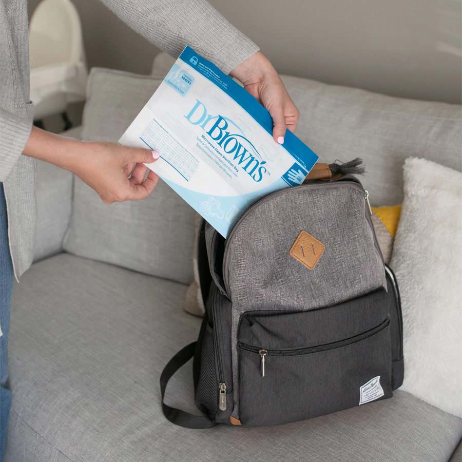 A person placing a Dr. Brown's sterilization pouch into an open grey backpack on a couch. The pouch is blue and white, while the backpack has multiple compartments and a zipper. The couch is light grey and has a white and yellow pillow beside the backpack.