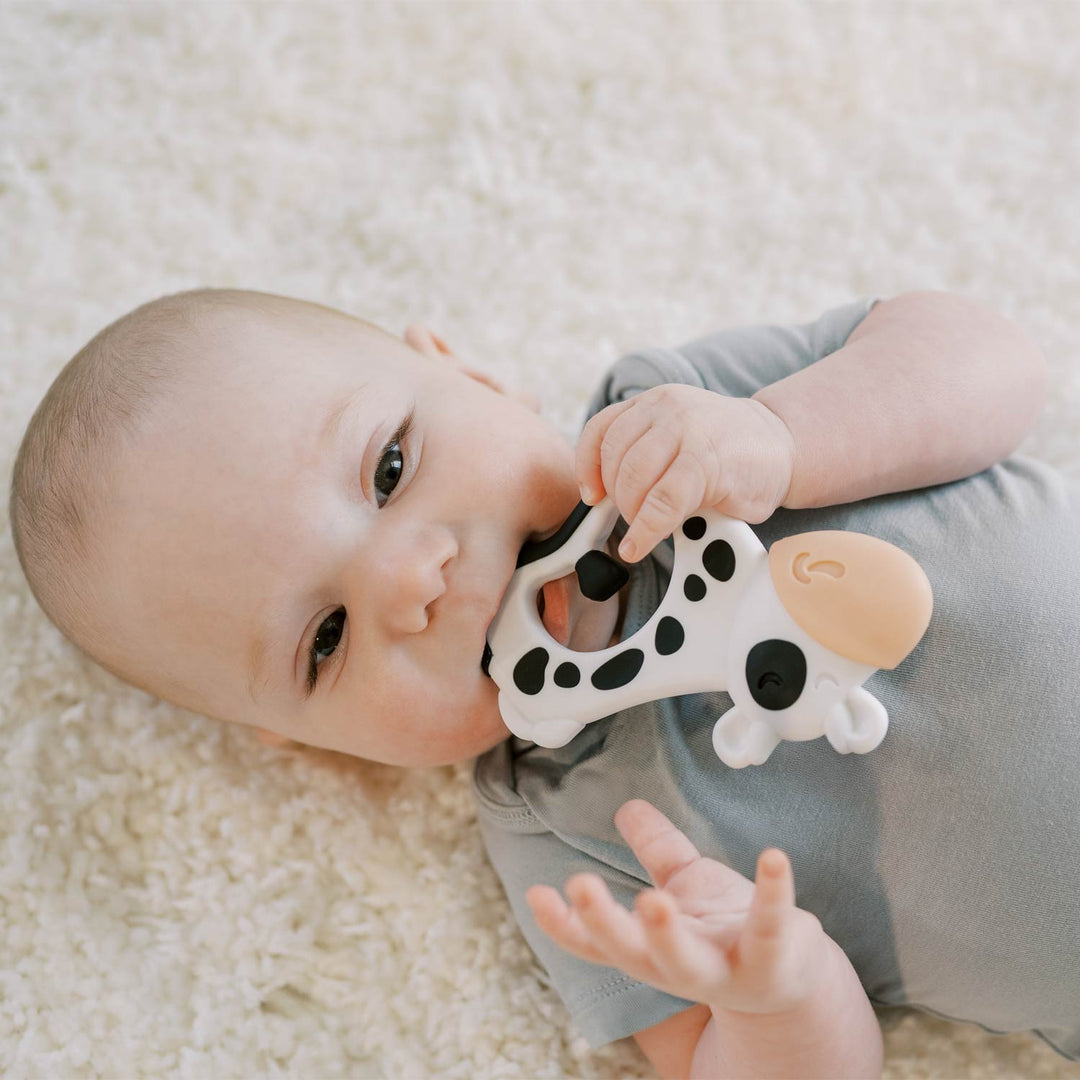 A baby lying on a soft cream rug, holding a white and black cow-shaped teething toy.