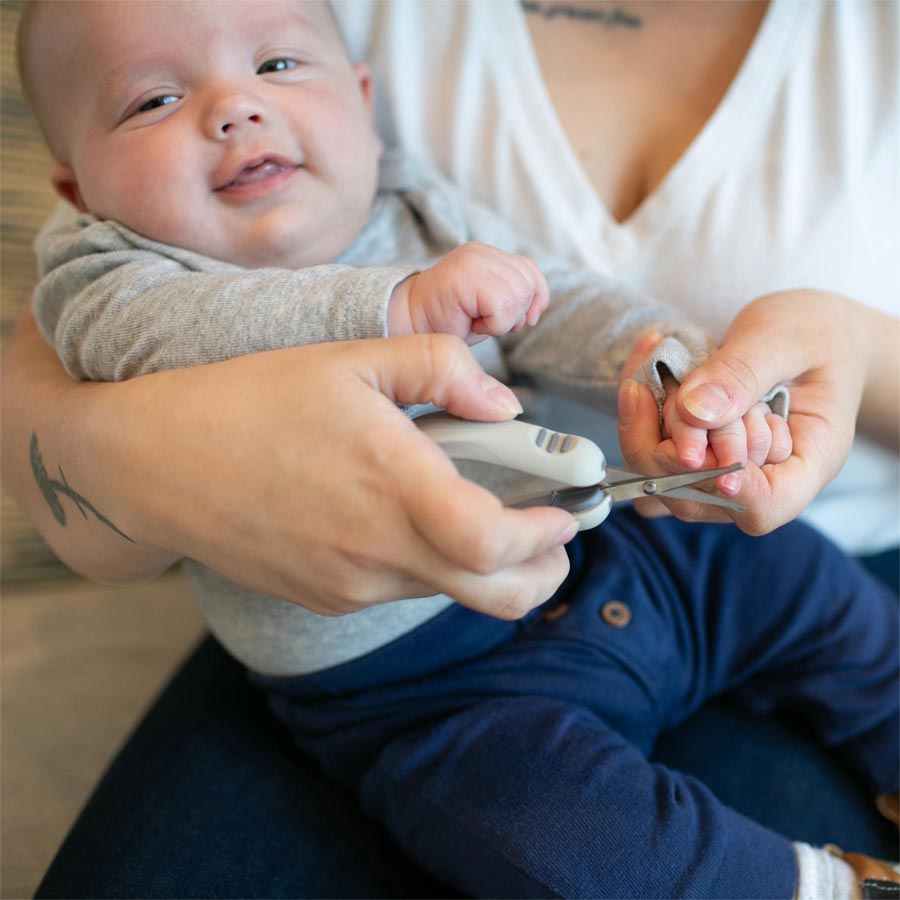 A caregiver gently clips a baby's fingernails while the baby, dressed in a gray top and blue pants, rests on their lap. The caregiver holds the baby's hand securely, ensuring a safe and comfortable nail trimming process.