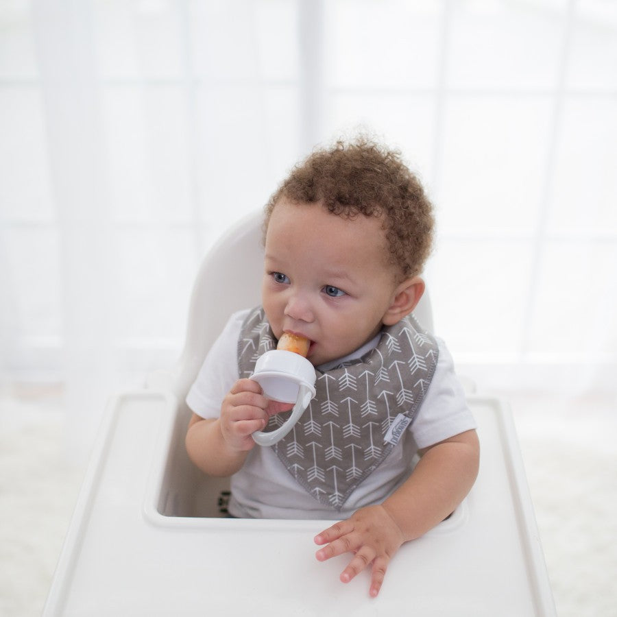 A baby in a high chair wearing a patterned bib is holding a baby food feeder and sucking on it. The background shows a softly lit window with sheer curtains.