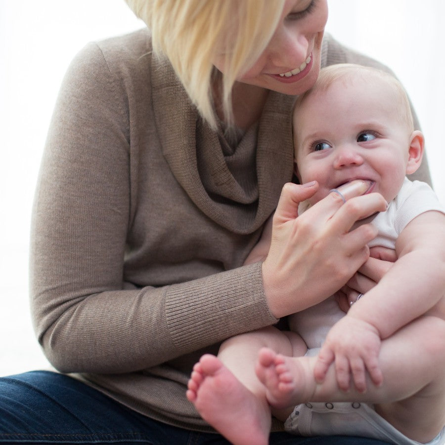 A smiling woman with short hair holds a happy baby in her lap. The woman is wearing a light brown sweater, and the baby is in a white onesie. They appear to be in a bright, softly lit room.