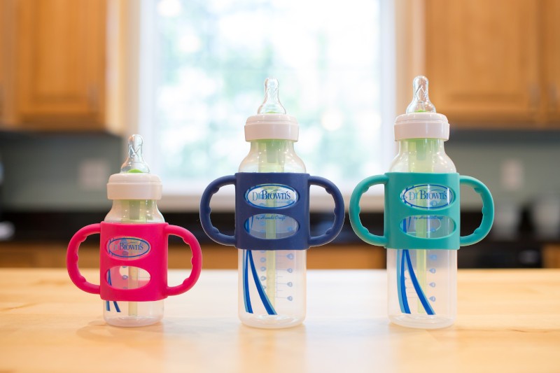 Three baby bottles of varying sizes are lined up on a wooden table. Each bottle has a different colored handle: pink, blue, and teal. They are against a blurred kitchen background with light coming through a window.