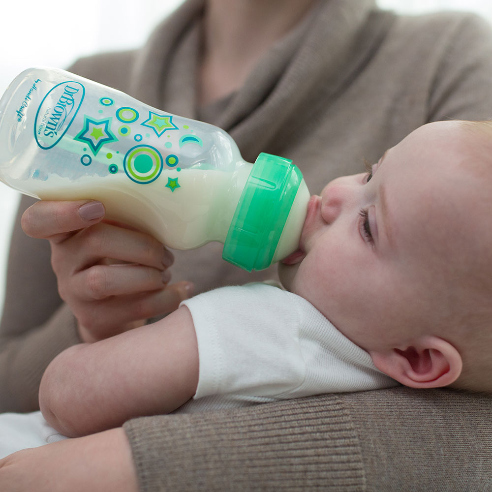 A person holds an infant who is drinking from a decorated baby bottle with a green cap. The baby is cradled in the person's arm. The background is softly blurred.