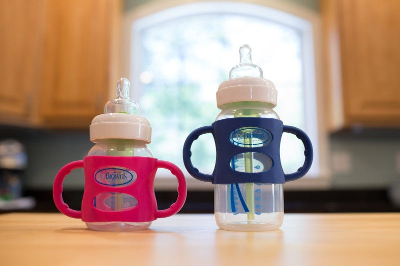 Two baby bottles with handles on a wooden table, one with a pink cover and the other with a blue cover. They are in a kitchen setting with blurred cabinets and a large window in the background.