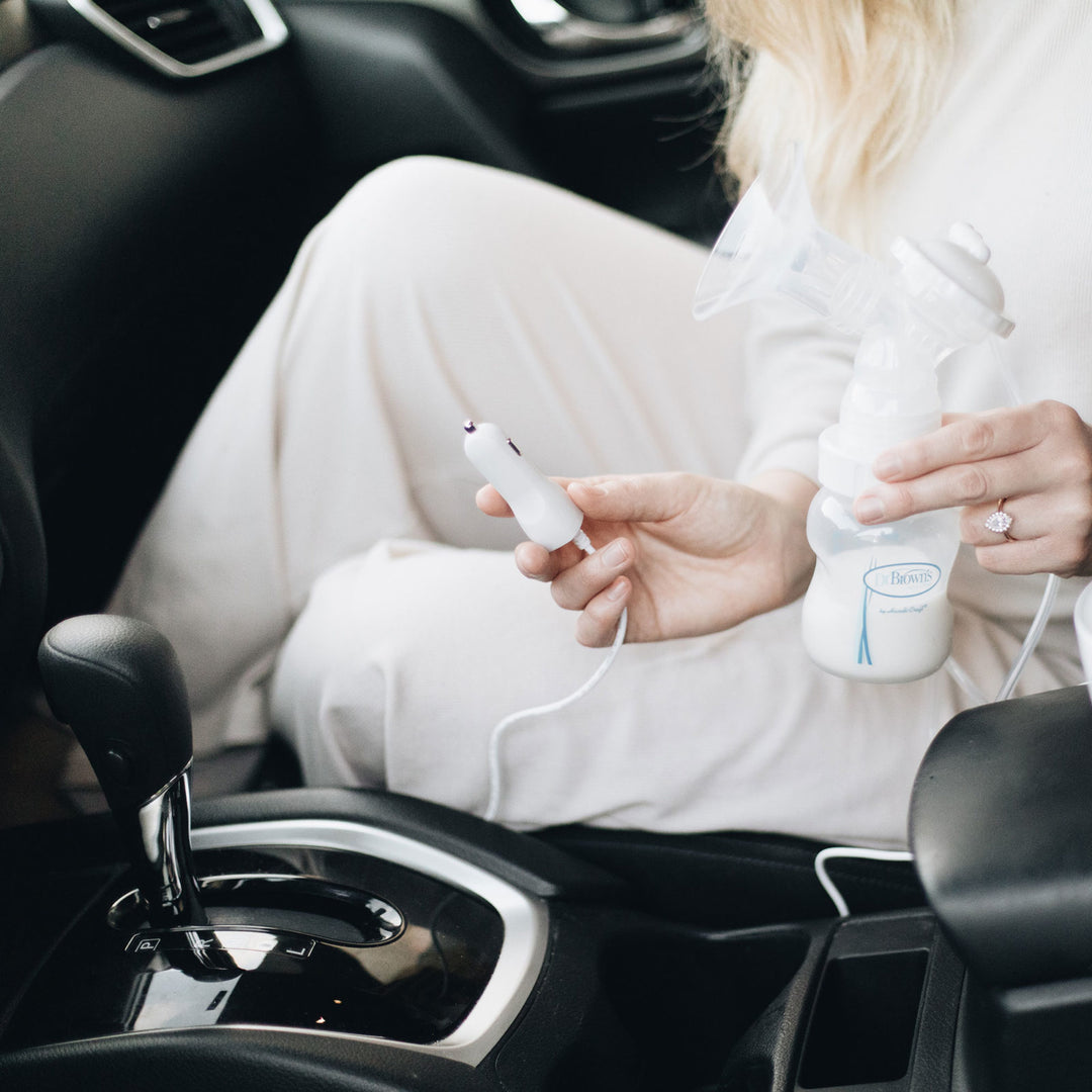 A person sitting in a car, holding a portable breast pump connected to a milk bottle. One hand adjusts the pump, while the other holds a car adapter plug. The person is wearing light-colored clothing.