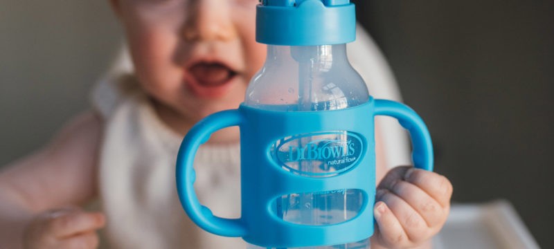 Baby in high chair holding silicone handle on blue sippy straw bottle