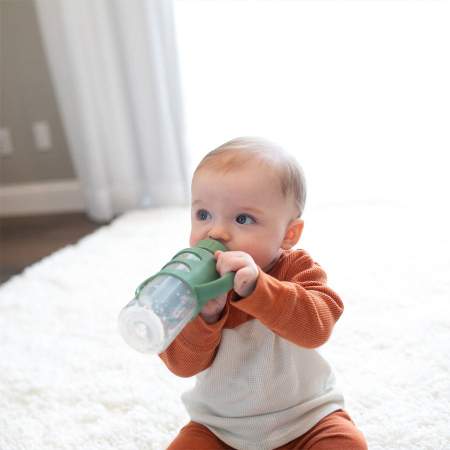 A baby in an orange and white outfit sits on a fluffy white rug, holding a green bottle with both hands. The background features soft lighting and curtains.