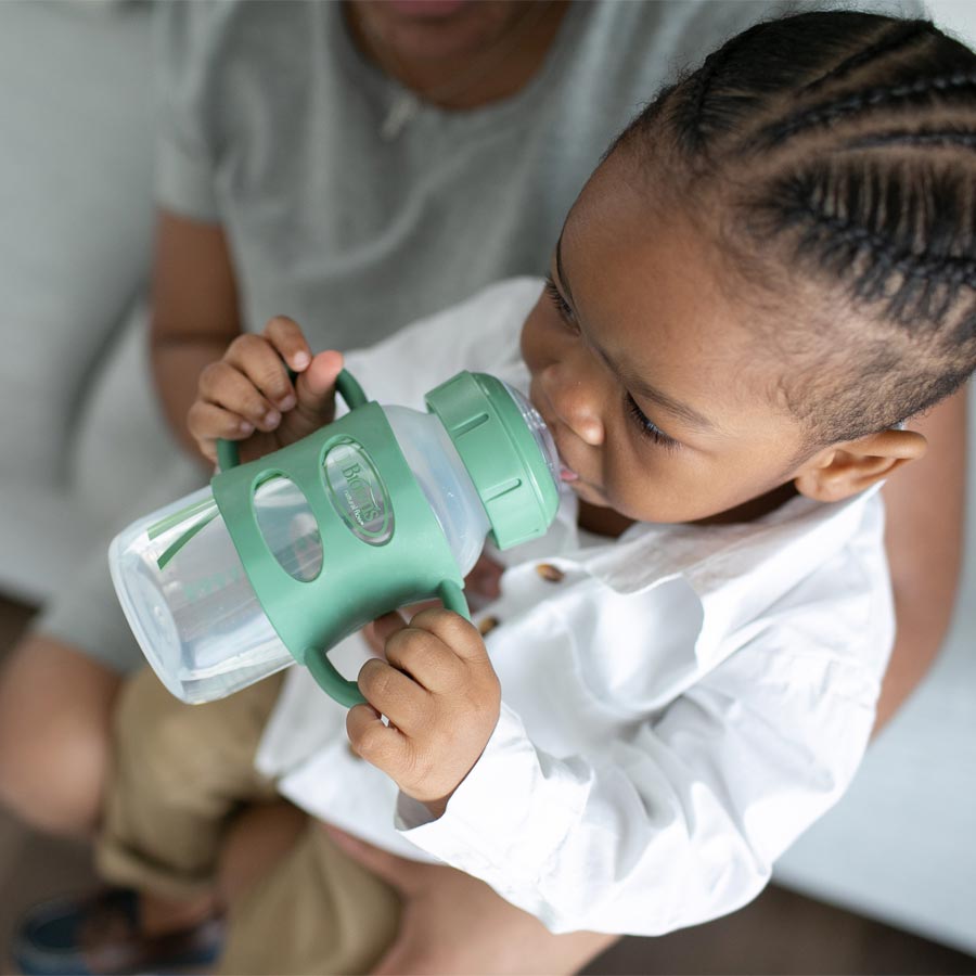 A toddler with braided hair, wearing a white shirt, drinks from a green-handled sippy cup while sitting on an adult's lap. The adult is partially visible and wears a gray shirt.