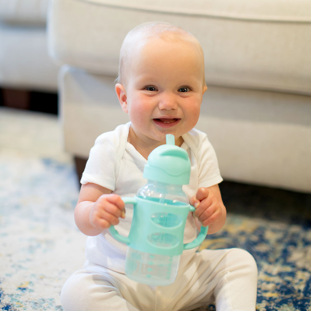 A smiling baby in a white outfit is sitting on a rug, holding a green sippy cup. A beige couch is in the background.