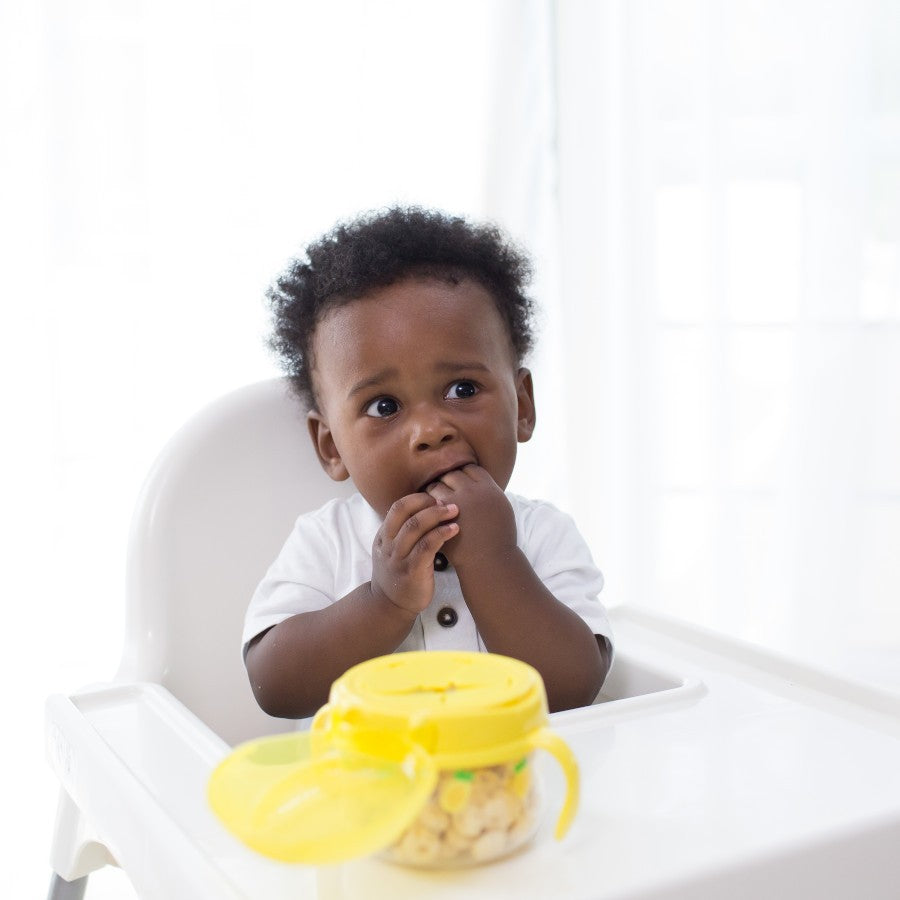 A baby with curly hair sits in a high chair, wearing a white shirt. They are eating snacks from a yellow container in front of them. The background is bright and softly lit.