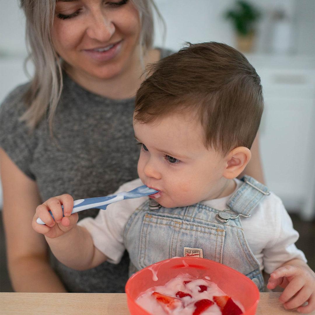 A baby wearing denim overalls sits at a table while a woman in a gray shirt smiles in the background. The baby holds a blue Soft-Tip Spoon and is eating from a red bowl containing yogurt and strawberries.