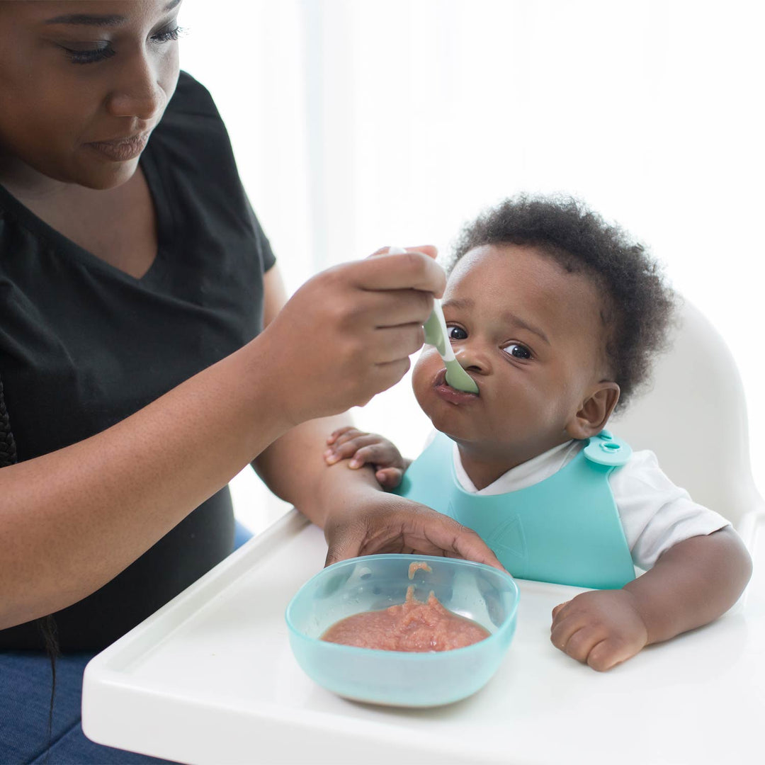 A woman feeds a baby using a light green Soft-Tip Spoon. The baby is wearing a light blue bib and sitting in a white high chair, with a small light blue bowl of food on the chair tray.