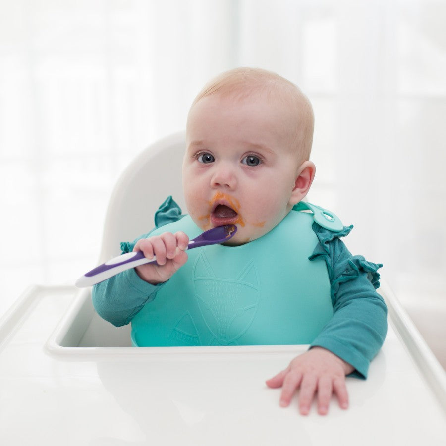 A baby sitting in a high chair, wearing a teal bib. The baby is holding a purple and white spoon, with some food around their mouth and on the spoon. The background is softly blurred.