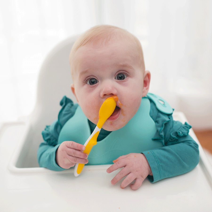 A baby wearing a teal bib sits in a highchair, holding a yellow spoon with food on it. The baby appears to be eating, with some food around the mouth. The background is bright and softly blurred.