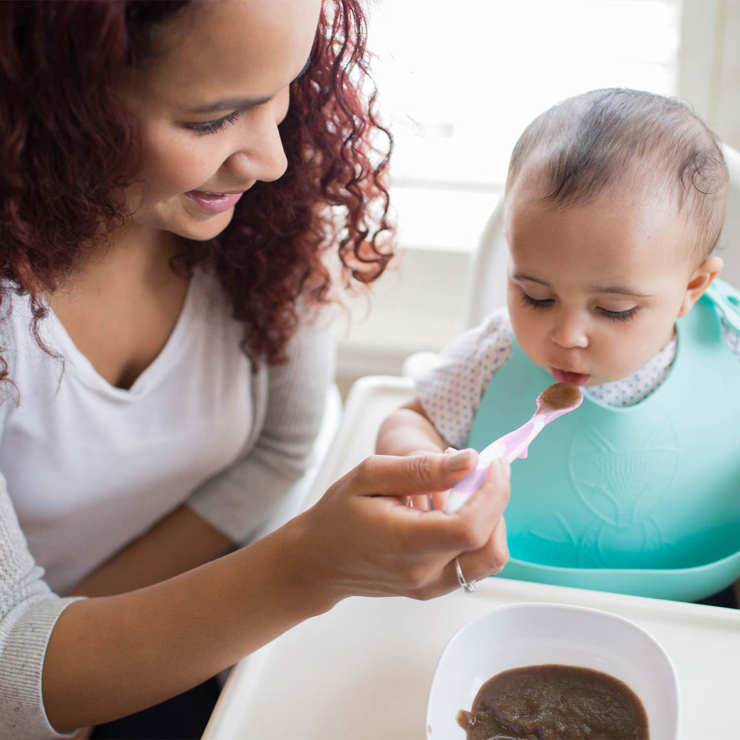 A woman feeding a baby with a pink spoon in a bright indoor setting. The baby is wearing a light blue bib and sitting in a high chair with a bowl of food in front.