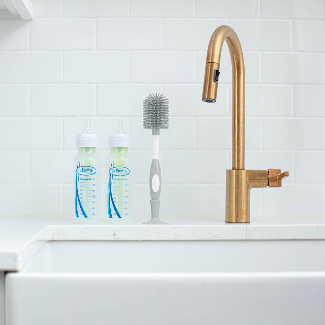 A modern kitchen sink scene with a tall brushed-gold faucet, two clear baby bottles with blue and white labels, and a gray-and-white bottle-cleaning brush against a white subway tile backsplash.