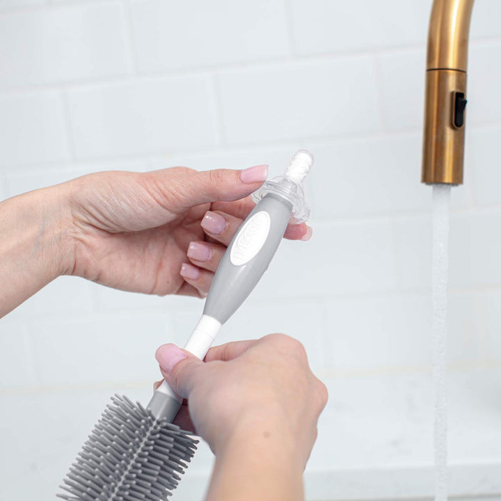 A person's hands holding a gray and white bottle brush and using it to clean a narrow nipple, positioned near a running faucet in a kitchen setting with white subway tiles.