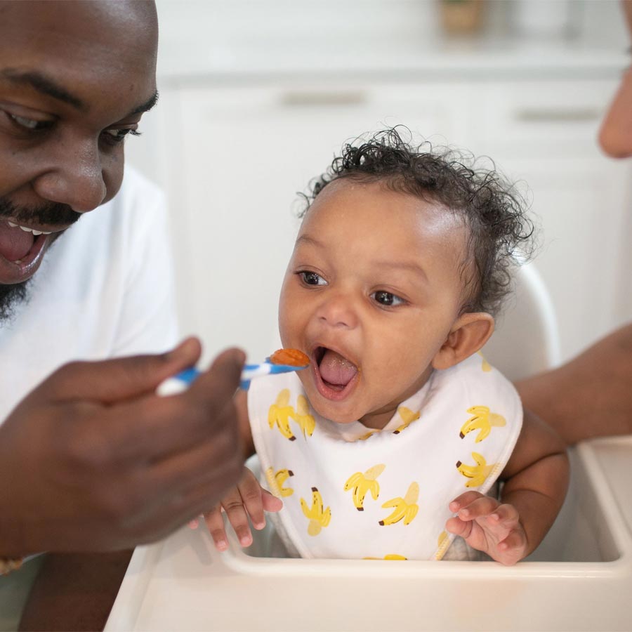 A baby wearing a bib with banana patterns sits in a high chair, opening their mouth as an adult feeds them with a spoon. Another person is partially visible, smiling at the baby. The setting appears to be a kitchen.