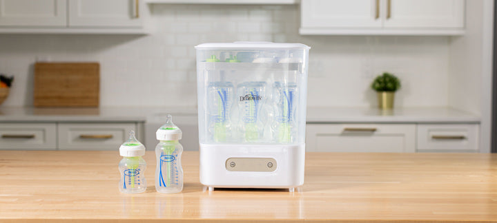A white and clear baby bottle sterilizer and dryer machine stands on a wooden countertop in a bright kitchen, with two baby bottles placed beside it.