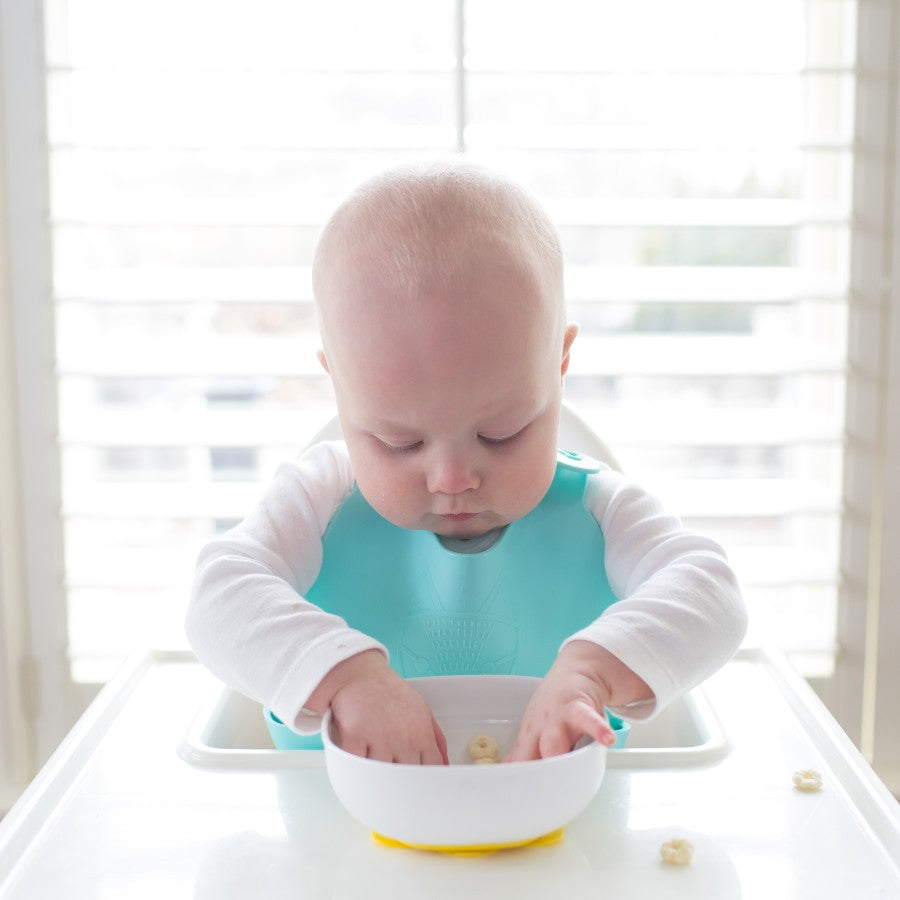 A baby with a blue bib sits in a high chair, focusing intently on a bowl of food, with hands inside the bowl. Soft light enters through the window blinds in the background.