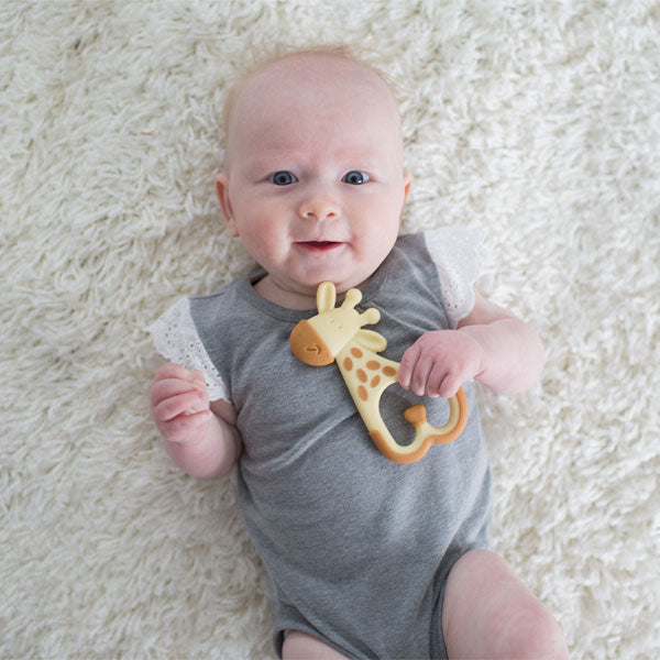 A baby in a gray onesie lies on a soft, white surface, holding a giraffe-shaped teething toy. The baby is looking up with a gentle expression.