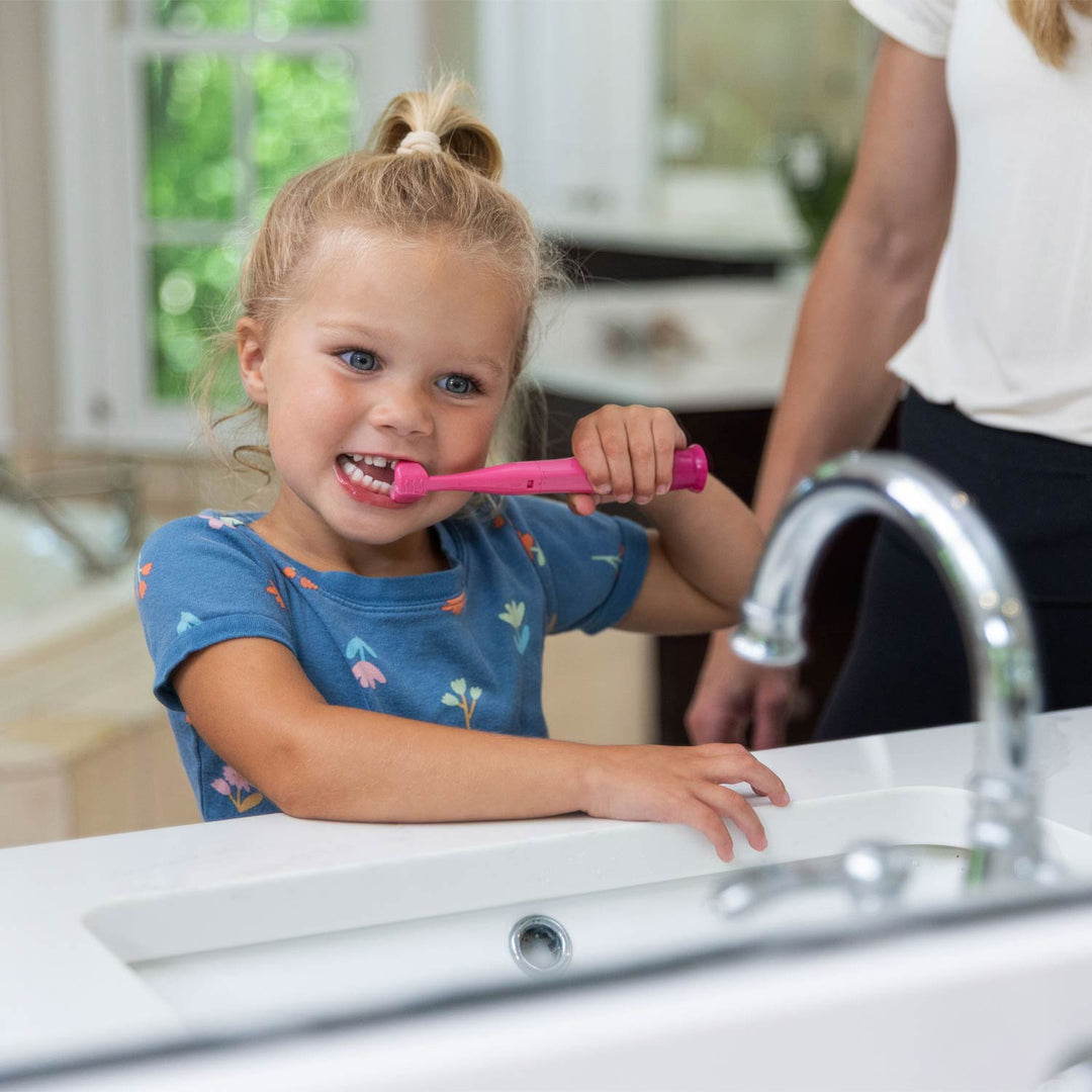 A young girl with blonde hair in a high ponytail smiles while brushing her teeth with a pink electric toothbrush at a white bathroom sink. An adult woman in a white shirt stands behind her.