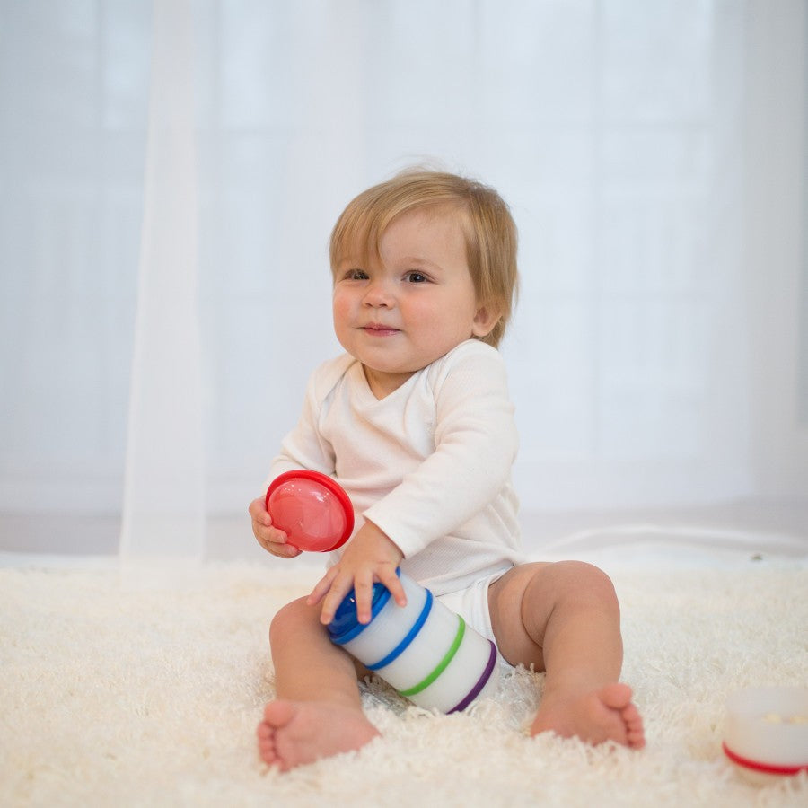 A baby with blonde hair, dressed in a white onesie, sits on a soft white carpet. The baby is holding and playing with colorful stacking cups, looking slightly to the side with a small smile. A light background enhances the cozy setting.