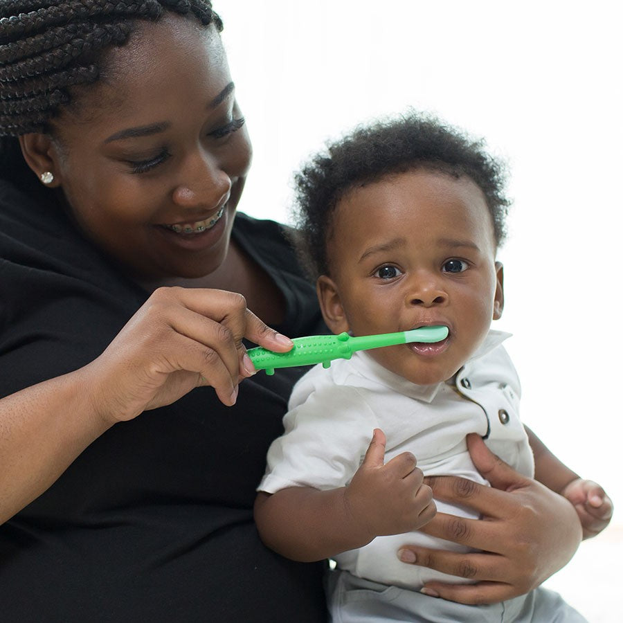 A woman holding a baby gently brushes the baby's teeth with a green toothbrush. The baby wears a white shirt and looks forward. The woman smiles warmly, wearing a black top with braided hair.