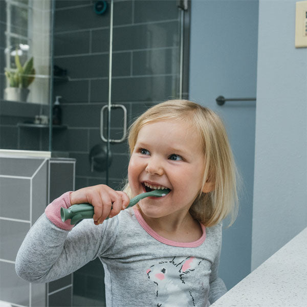 A smiling child with blonde hair is brushing their teeth in a bathroom. The child is wearing a gray pajama top with a bunny design. There's a modern glass shower in the background and a potted plant on a shelf.