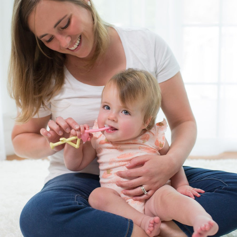 A smiling woman sits on the floor, holding a toddler in her lap. The toddler is wearing a pink onesie and is playfully chewing on a plastic toy. Both appear happy in a bright room with natural light filtering through large windows.