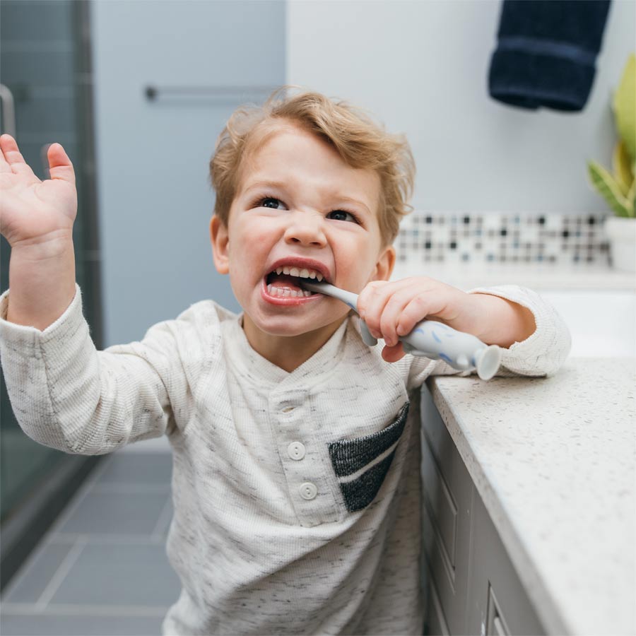 A young child in a white shirt energetically brushes their teeth in a bathroom. The child is making a playful expression, standing next to a countertop with a plant and bathroom accessories in the background.