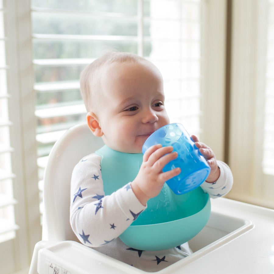 A baby sitting in a high chair, wearing a bib with blue accents and a star-patterned outfit, is holding and drinking from a light blue cup. The background features bright windows with white blinds.