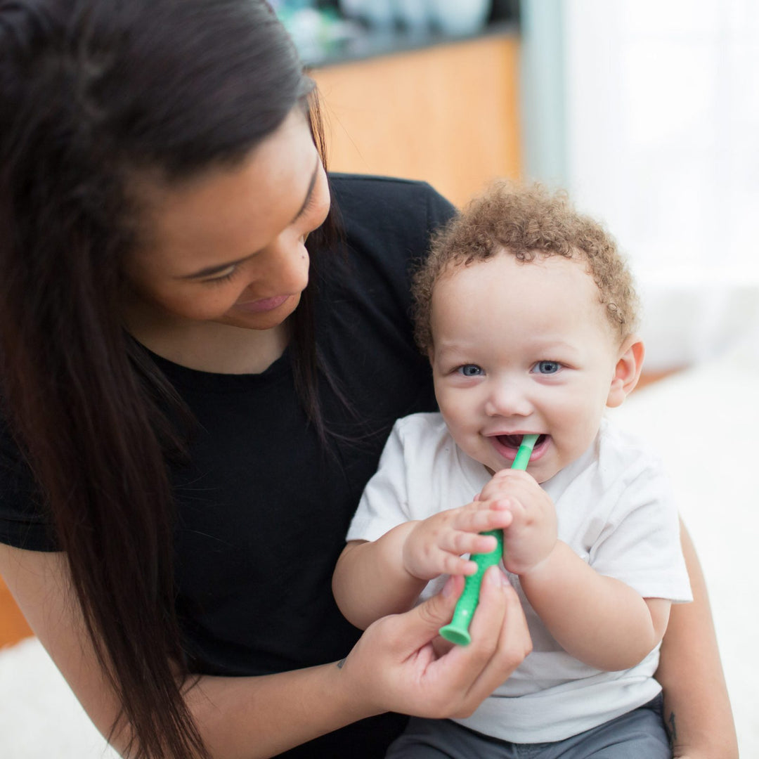 A woman lovingly holds a smiling baby on her lap. She helps the baby brush his teeth with a green toothbrush. The background is softly blurred, creating a warm and cozy atmosphere.