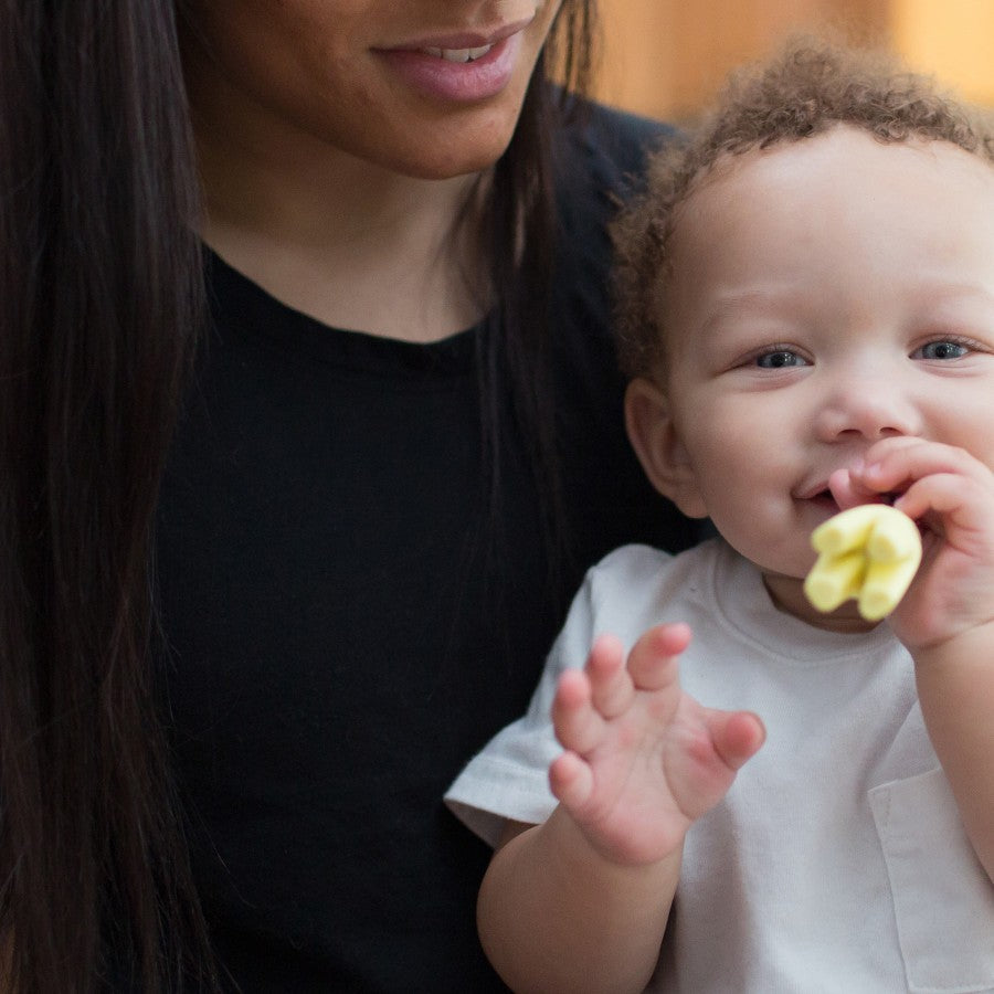 A baby in a white shirt chewing on a yellow toy is held by an adult. The adult's face is partially shown, with long dark hair and wearing a black shirt. The background is softly blurred, creating a warm and intimate atmosphere.